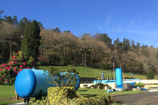 Imagen de tuberías y depósitos pintados de azul en un entorno verde con arbolado.